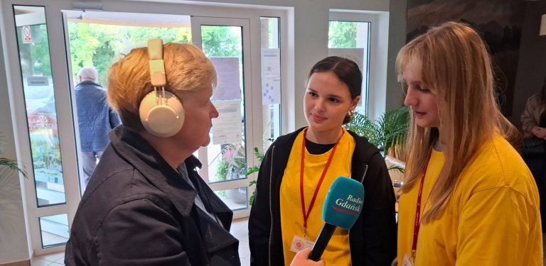 Dziennikarka Radia Gdańsk Anna Rębas w słuchawkach na głowie. Trzyma mikrofon i rozmawia z dwiema młodymi wolontariuszkami ubranymi w żółte koszulki Caritas. 30-lecie działalności Centrum Pomocowego Caritas im. św. Faustyny w Rumi
