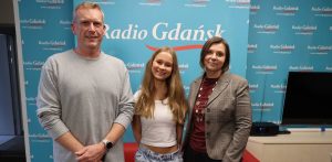 Trzy uśmiechnięte osoby w studiu Radia Gdańsk na tle niebieskiej ścianki z logo stacji. Od lewej stoją: Krzysztof Kowalski w jasnoszarym swetrze, wolontariuszka Karolina Mamprejew w białej bluzce oraz Marta Czyż w ciemnym żakiecie