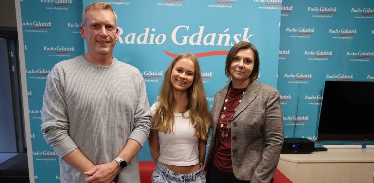 Trzy uśmiechnięte osoby w studiu Radia Gdańsk na tle niebieskiej ścianki z logo stacji. Od lewej stoją: Krzysztof Kowalski w jasnoszarym swetrze, wolontariuszka Karolina Mamprejew w białej bluzce oraz Marta Czyż w ciemnym żakiecie