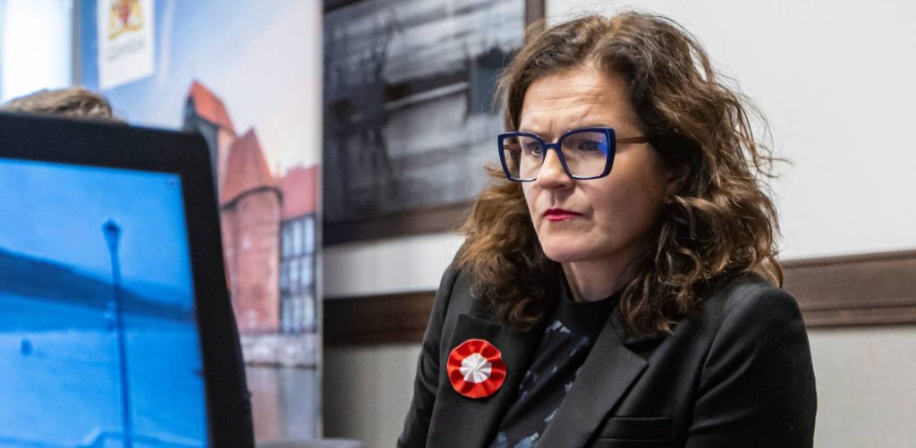 Prezydent Gdańska Aleksandra Dulkiewicz w wyrazistych okularach i ciemnym żakiecie z wpiętą biało-czerwoną rozetką, ze skupionym, poważnym wyrazem twarzy