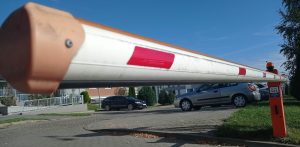 Opuszczony biało-czerwony szlaban wjazdowy na parking zamkniętego osiedla mieszkaniowego, w tle zaparkowane samochody i bloki mieszkalne