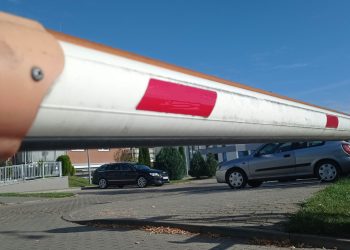 Opuszczony biało-czerwony szlaban wjazdowy na parking zamkniętego osiedla mieszkaniowego, w tle zaparkowane samochody i bloki mieszkalne