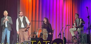 Zespół muzyczny na scenie: klarnecista, skrzypek, śpiewająca kobieta grająca na tradycyjnym bębnie oraz akordeonista. W tle kurtyna oświetlona na fioletowo i pomarańczowo. Kapela Begebenheit na scenie podczas festiwalu Cassubia Cantat