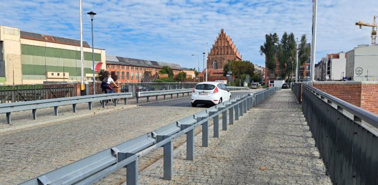 Most w Elblągu. Biały samochód osobowy oraz rowerzysta na moście wyłożonym kostką brukową, oddzielonym stalowymi barierami od chodnika. W tle widoczny zabytkowy, ceglany kościół w stylu gotyckim, zabudowania przemysłowe oraz błękitne niebo z chmurami