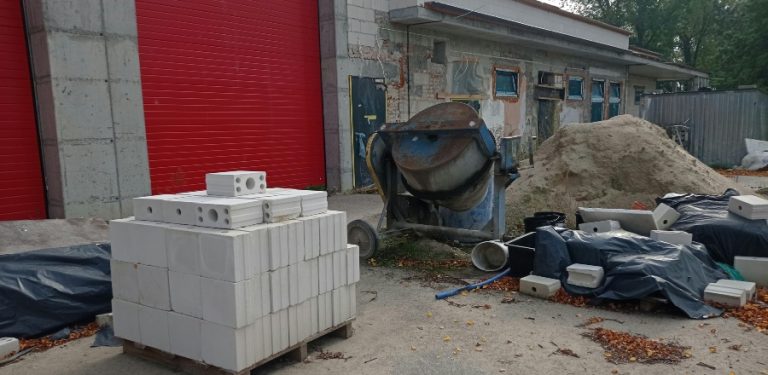 Plac budowy przed przebudowywaną siedzibą Gminnego Ośrodka Kultury i Sportu oraz remizy OSP w Lichnowach. Na pierwszym planie widać paletę z białymi bloczkami, stojącą obok betoniarkę, stertę piasku oraz materiały budowlane przykryte czarną folią.