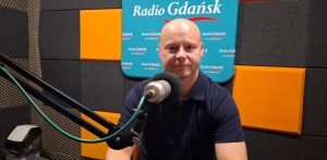 Mężczyzna (Łukasz Iwaszkiewicz) w ciemnej koszulce polo siedzi przed mikrofonem w studiu nagraniowym, w tle widoczne logo Radia Gdańsk na turkusowo-pomarańczowej ściance akustycznej
