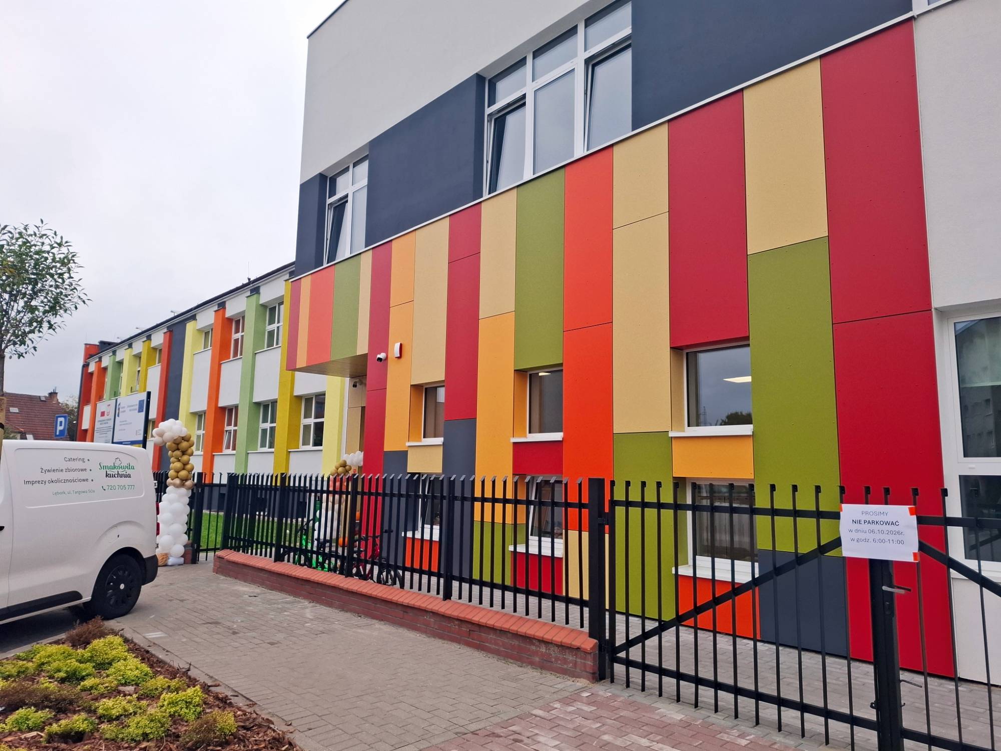 Nowoczesny, dwukondygnacyjny budynek żłobka i przedszkola w Lęborku z kolorową elewacją w pionowe pasy (czerwone, pomarańczowe, zielone i żółte). Przed wejściem widoczne czarne ogrodzenie oraz brama udekorowana girlandami z balonów
