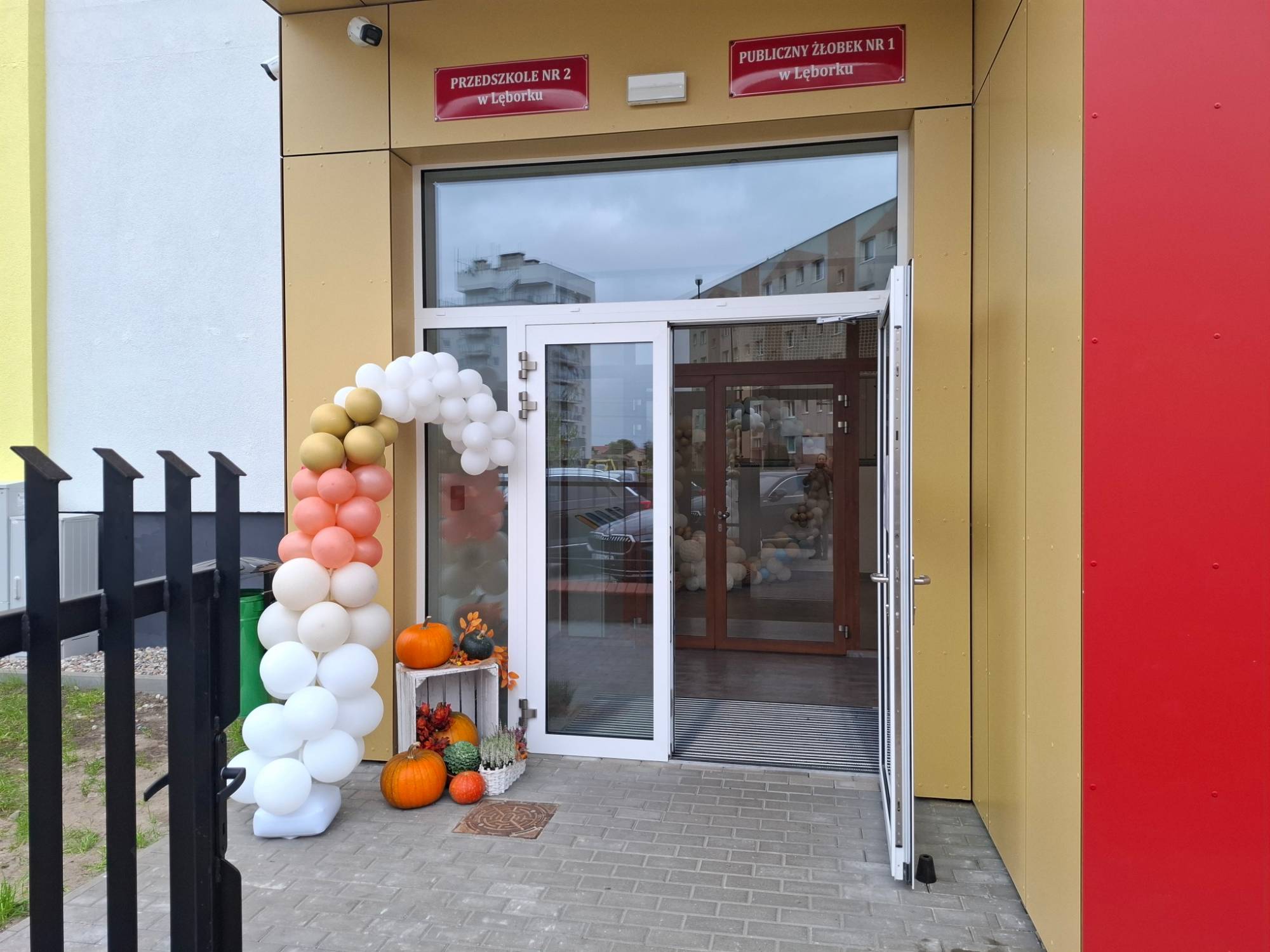 Wejście do budynku z dwiema czerwonymi tablicami: "Przedszkole nr 2 w Lęborku" oraz "Publiczny Żłobek nr 1 w Lęborku". Przed przeszklonymi drzwiami stoją dekoracje: łuk z pastelowych balonów oraz jesienna kompozycja z dyń i wrzosów