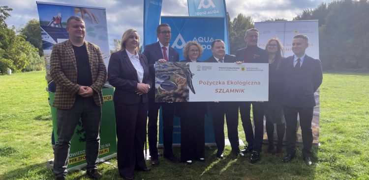 Przedstawiciele władz Sopotu, samorządu województwa pomorskiego oraz instytucji finansujących stoją w plenerze na trawniku i trzymają pamiątkową tablicę z napisem "Pożyczka Ekologiczna SZLAMNIK". W tle widoczne są banery promocyjne AQUA Sopot i Pomorskiego Funduszu Rozwoju