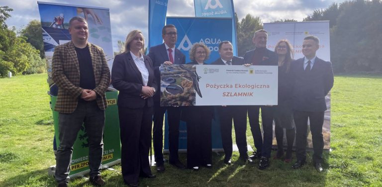 Przedstawiciele władz Sopotu, samorządu województwa pomorskiego oraz instytucji finansujących stoją w plenerze na trawniku i trzymają pamiątkową tablicę z napisem "Pożyczka Ekologiczna SZLAMNIK". W tle widoczne są banery promocyjne AQUA Sopot i Pomorskiego Funduszu Rozwoju