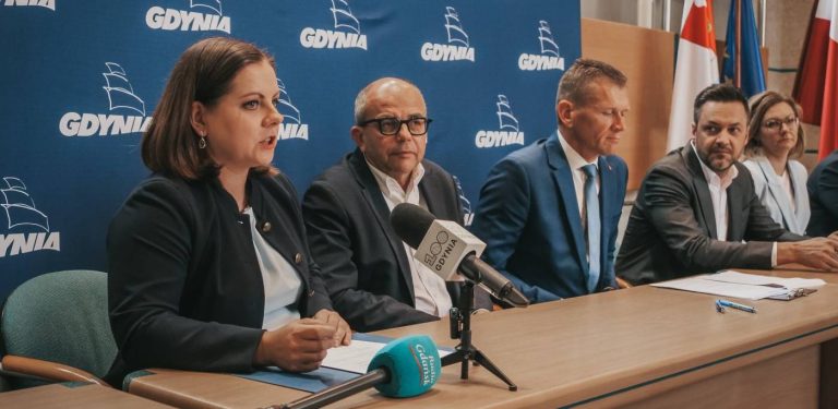 Konferencja prasowa w Gdyni. Przy stole konferencyjnym na tle niebieskiej ścianki z herbem i logo miasta siedzą przedstawiciele władz i zaproszeni goście. Na pierwszym planie po lewej stronie przemawia prezydent Gdyni Aleksandra Kosiorek, przed którą na blacie leżą dokumenty oraz mikrofony stacji radiowych.