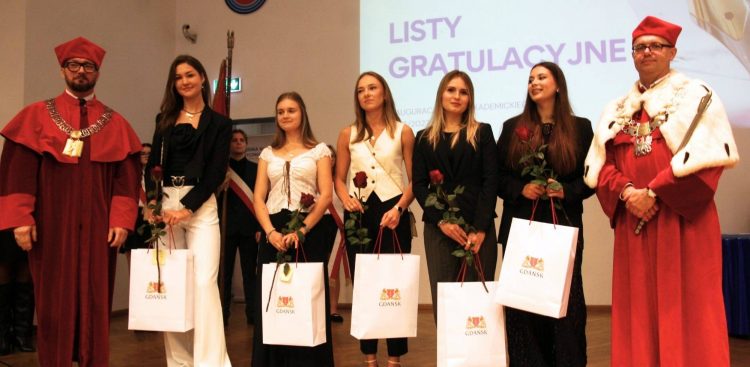 Władze AWFiS w Gdańsku w czerwonych togach i łańcuchach rektorskich stoją po obu stronach grupy pięciu studentek trzymających czerwone róże oraz białe torby z herbem Gdańska. W tle widoczny jest ekran z napisem "Listy gratulacyjne"