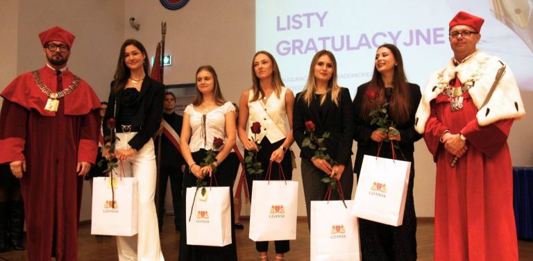 Władze AWFiS w Gdańsku w czerwonych togach i łańcuchach rektorskich stoją po obu stronach grupy pięciu studentek trzymających czerwone róże oraz białe torby z herbem Gdańska. W tle widoczny jest ekran z napisem "Listy gratulacyjne"