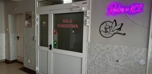 Białe, przeszklone drzwi do sali porodowej z różowym napisem "SALA PORODOWA". Po prawej stronie na szarej ścianie widnieje świecący na różowo neon z napisem "Rodzę w KCZ" oraz metalowa ozdoba przedstawiająca lecącego bociana z noworodkiem w zawiniątku