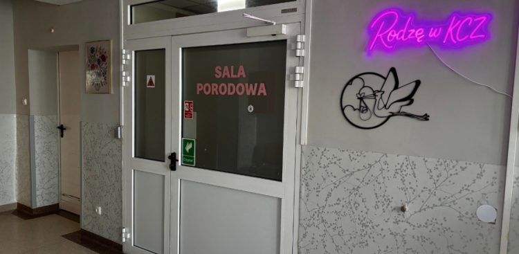 Białe, przeszklone drzwi do sali porodowej z różowym napisem "SALA PORODOWA". Po prawej stronie na szarej ścianie widnieje świecący na różowo neon z napisem "Rodzę w KCZ" oraz metalowa ozdoba przedstawiająca lecącego bociana z noworodkiem w zawiniątku