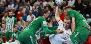 Piłkarz ręczny w białym stroju Wybrzeża Gdańsk, trzymający piłkę w uniesionej dłoni, jest dynamicznie powstrzymywany przez dwóch obrońców macedońskiego Eurofarmu Pelister w zielonych koszulkach