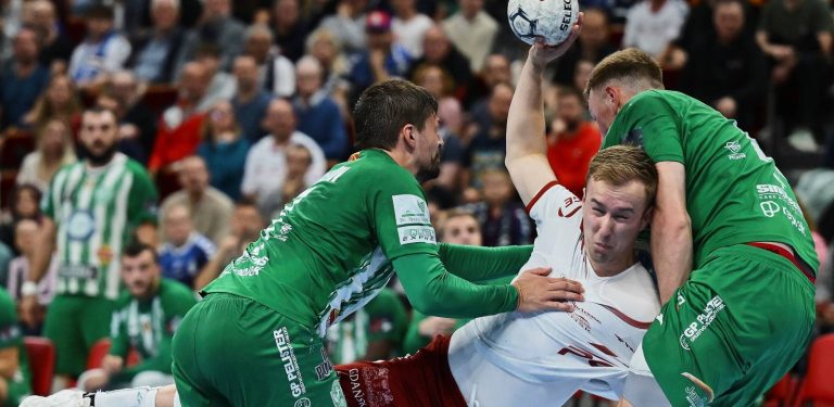Piłkarz ręczny w białym stroju Wybrzeża Gdańsk, trzymający piłkę w uniesionej dłoni, jest dynamicznie powstrzymywany przez dwóch obrońców macedońskiego Eurofarmu Pelister w zielonych koszulkach