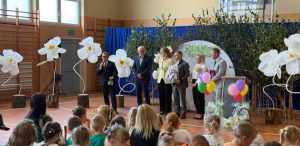 Żłobek w Gołubiu. Uroczystość w sali gimnastycznej udekorowanej wielkimi białymi kwiatami i balonami. Na scenie przemawia mężczyzna z mikrofonem w otoczeniu oficjeli. Na pierwszym planie widoczna jest widownia złożona z siedzących dzieci i opiekunów
