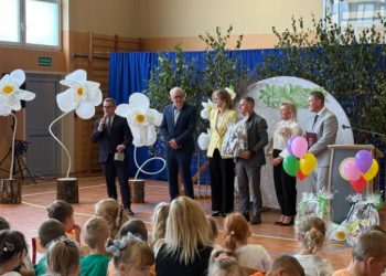 Żłobek w Gołubiu. Uroczystość w sali gimnastycznej udekorowanej wielkimi białymi kwiatami i balonami. Na scenie przemawia mężczyzna z mikrofonem w otoczeniu oficjeli. Na pierwszym planie widoczna jest widownia złożona z siedzących dzieci i opiekunów