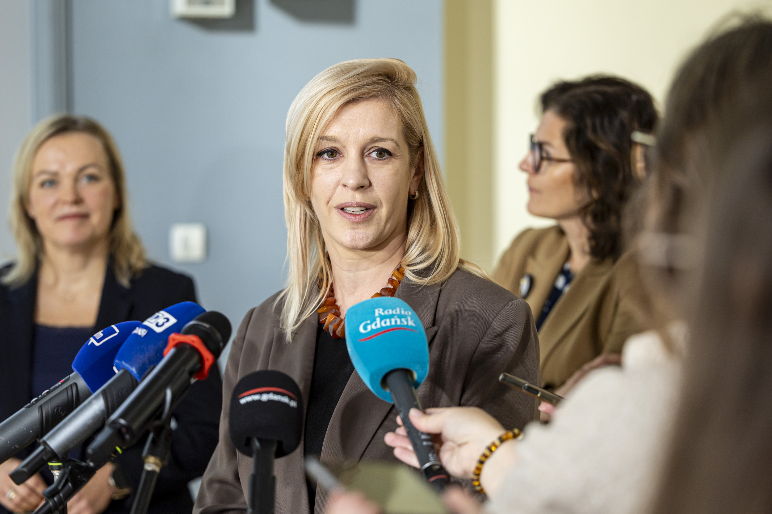 Zastępczyni prezydenta Gdańska Emilia Lodzińska, blondynka w ciemnej marynarce i naszyjniku, przemawia do mikrofonów dziennikarzy na konferencji prasowej. W tle po prawej stronie widać prezydent Gdańska Aleksandrę Dulkiewicz
