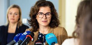 Prezydent Gdańska Aleksandra Dulkiewicz w okularach i beżowym żakiecie przemawia do mikrofonów dziennikarzy podczas konferencji prasowej. W tle widoczna jest kobieta