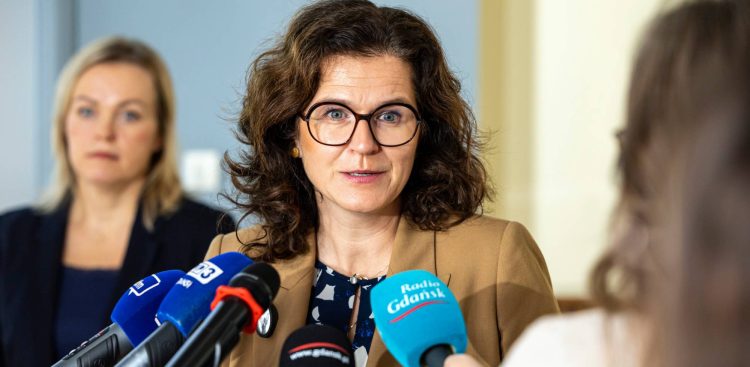 Prezydent Gdańska Aleksandra Dulkiewicz w okularach i beżowym żakiecie przemawia do mikrofonów dziennikarzy podczas konferencji prasowej. W tle widoczna jest kobieta