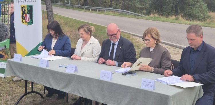 Pięcioro samorządowców i przedstawicieli inwestycji siedzi na świeżym powietrzu przy stole nakrytym obrusem i podpisuje dokumenty przy drodze. W tle widoczny jest baner z herbem powiatu kościerskiego oraz asfaltowa jezdnia.