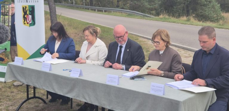 Pięcioro samorządowców i przedstawicieli inwestycji siedzi na świeżym powietrzu przy stole nakrytym obrusem i podpisuje dokumenty przy drodze. W tle widoczny jest baner z herbem powiatu kościerskiego oraz asfaltowa jezdnia.