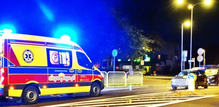 Karetka pogotowia ratunkowego na sygnale stojąca w nocy na jezdni przy oznakowanym przejściu dla pieszych; w tle widoczne zatrzymane samochody osobowe i latarnie uliczne